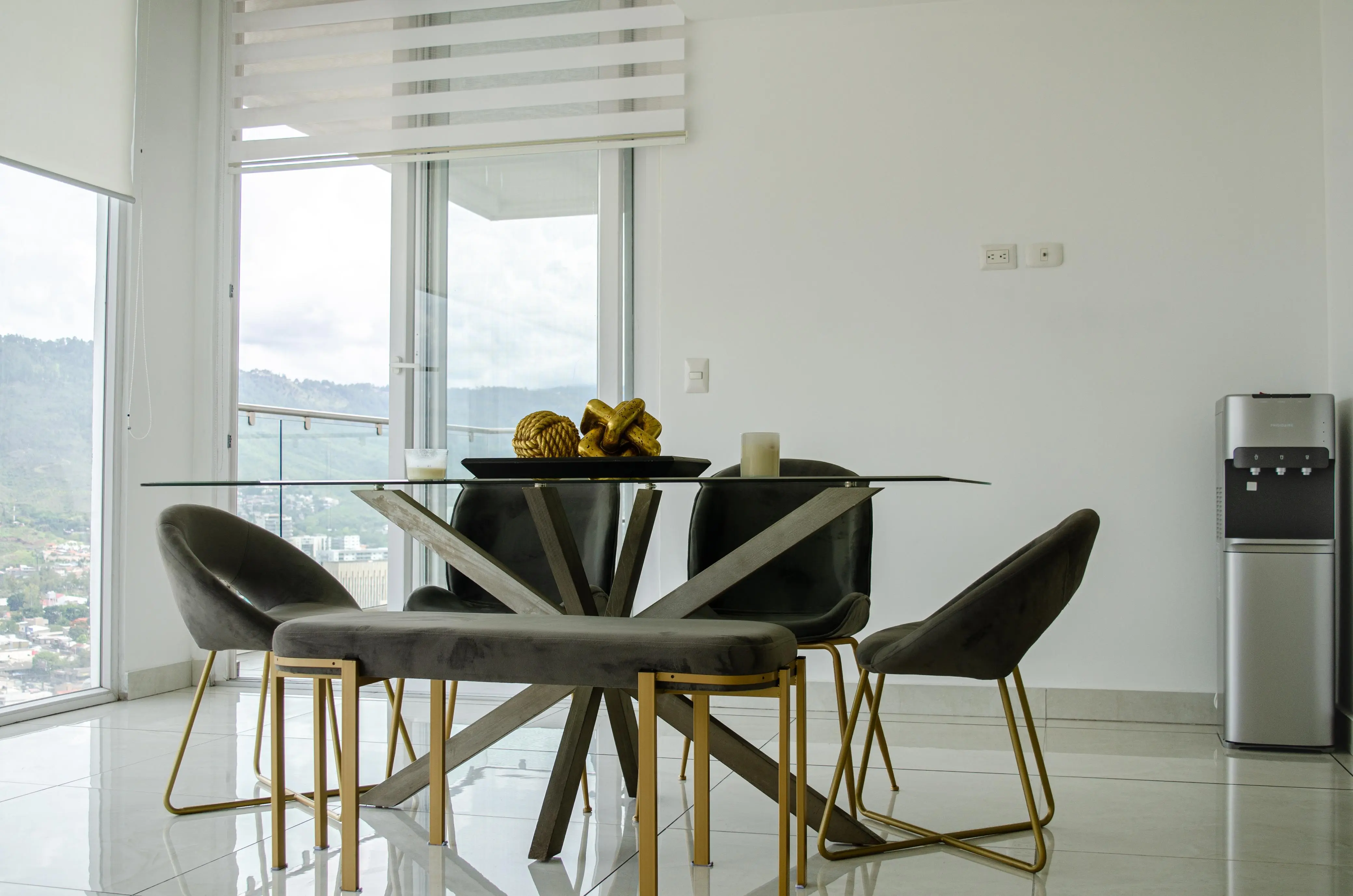Comedor de vidrio con sillas de terciopelo gris y patas de metal, junto a la puerta del balcón.