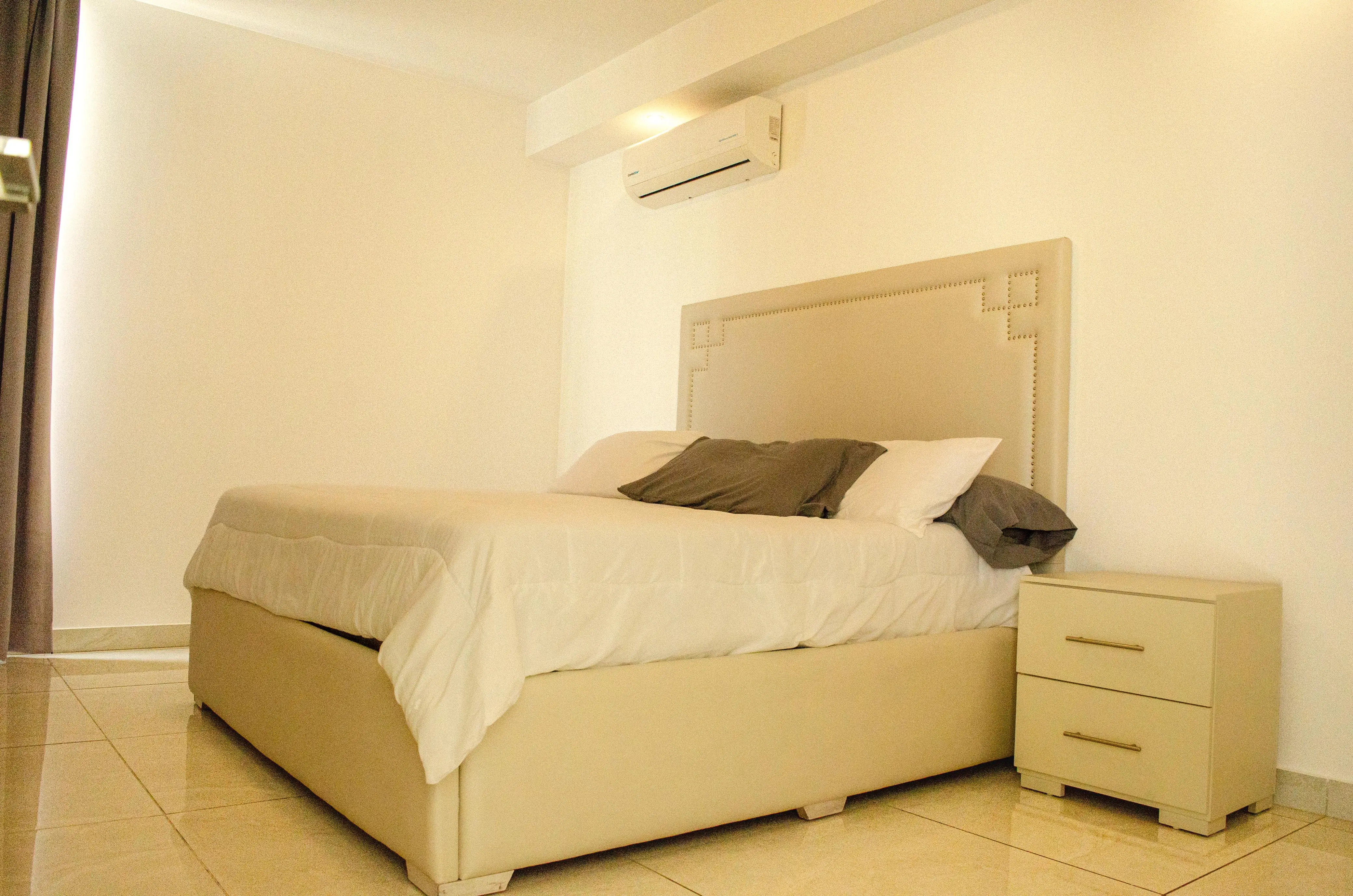 Habitación con cama vestida de blanco, cabecera beige con tachuelas, buró y aire acondicionado.