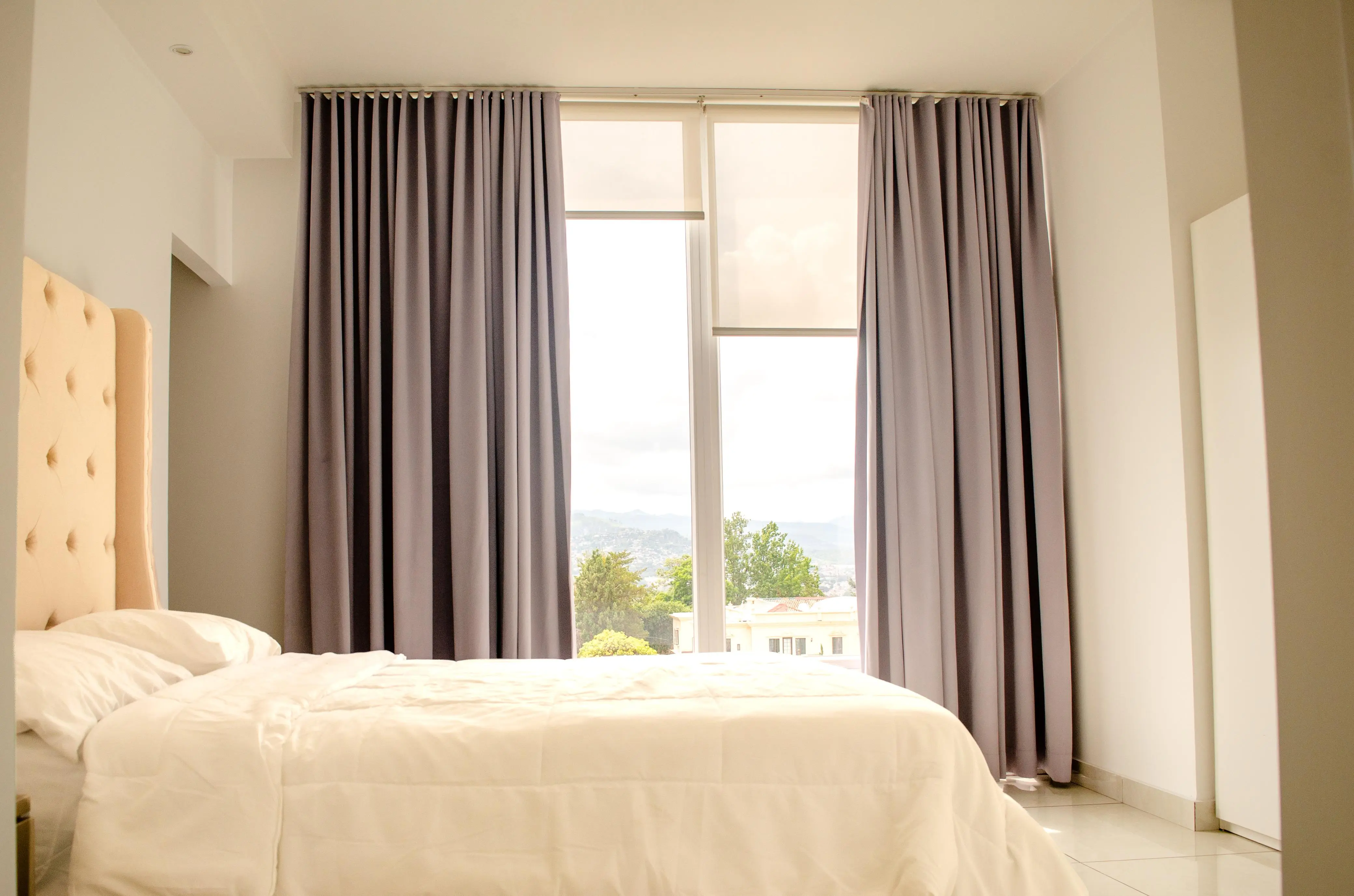 Habitación con cama vestida de blanco, cabecera capitoné y cortinas grises abiertas hacia la ventana y la ciudad.